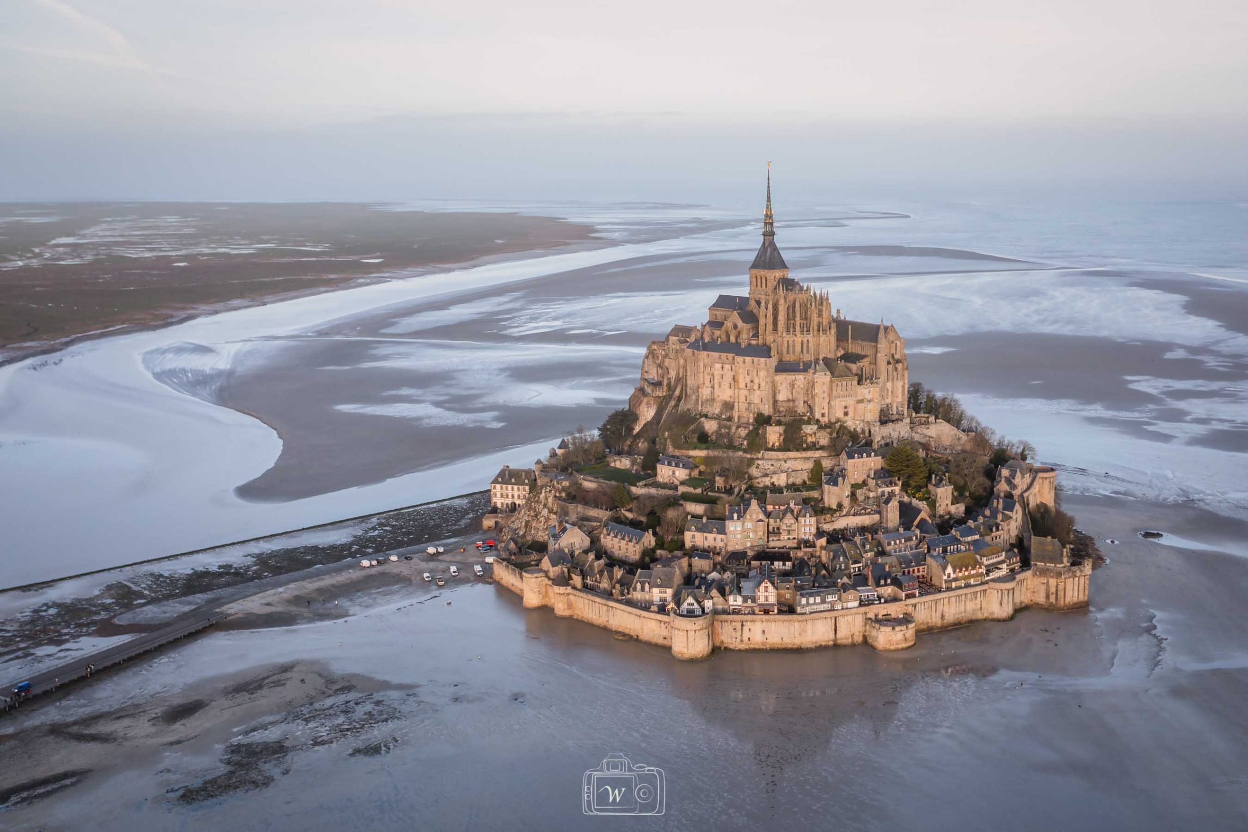 Vue aérienne du Mont-Saint-Michel à marée basse, avec l’abbaye surplombant le village et les vastes étendues de sables en arrière-plan, baignées dans une lumière douce et diffuse.