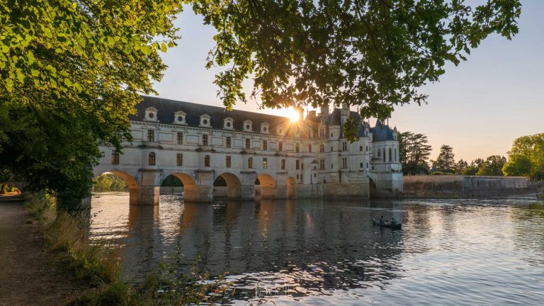 Vue en contre-jour de la galerie et des arches du château de Chenonceau enjambant le Cher au coucher du soleil, encadrée par le feuillage des arbres.