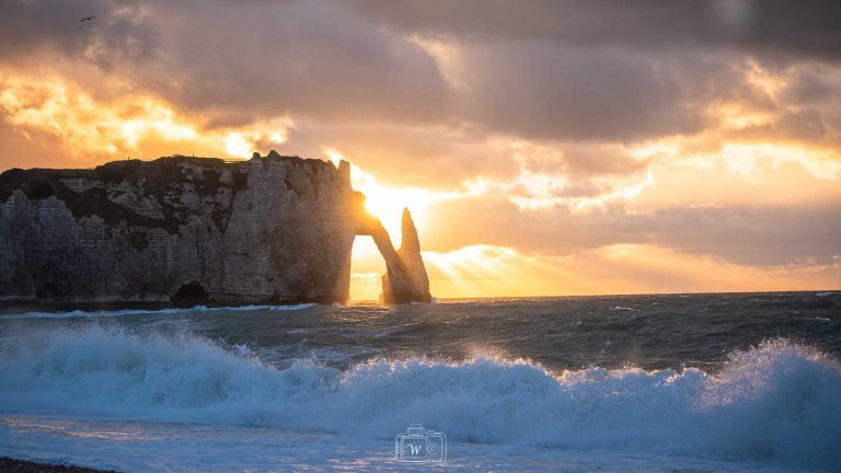 Coucher de soleil derrière l’aiguille et l’arche d’Étretat, illuminant les falaises crayeuses et les vagues de la Manche sous un ciel dramatique.