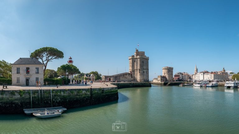 Vue panoramique sur le chenal d'entrée du Vieux-Port de La Rochelle, avec le quai de la Fouras au premier plan, la tour médiévale Saint-Nicolas en pierre calcaire et la tour de la Chaîne sous un ciel bleu limpide.