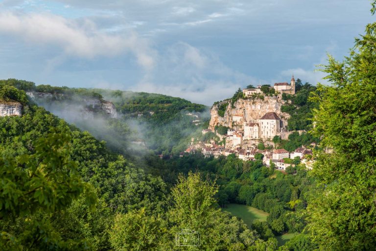Vue panoramique de Rocamadour au petit matin, avec les brumes matinales s'élevant entre les vallées boisées et le village perché sur sa falaise.