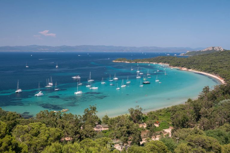 Vue panoramique en hauteur de la baie de la Courtade sur l'île de Porquerolles. L'eau dégrade du turquoise clair au bleu profond. De nombreux voiliers et catamarans sont au mouillage dans cette anse calme. Une forêt de pins borde la plage de sable blanc, avec les reliefs de la côte varoise visibles à l'horizon.