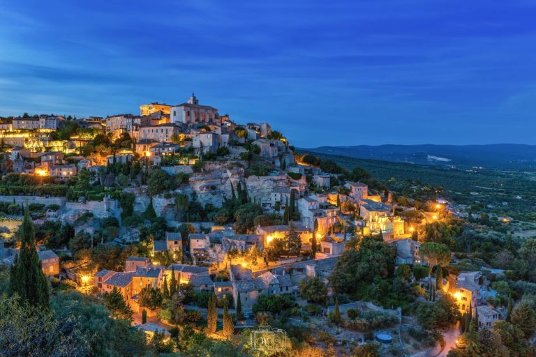 Vue panoramique à l'heure bleue sur le village perché de Gordes, aux maisons en pierre calcaire étagées à flanc de falaise et illuminées de teintes dorées face à la plaine du Luberon.