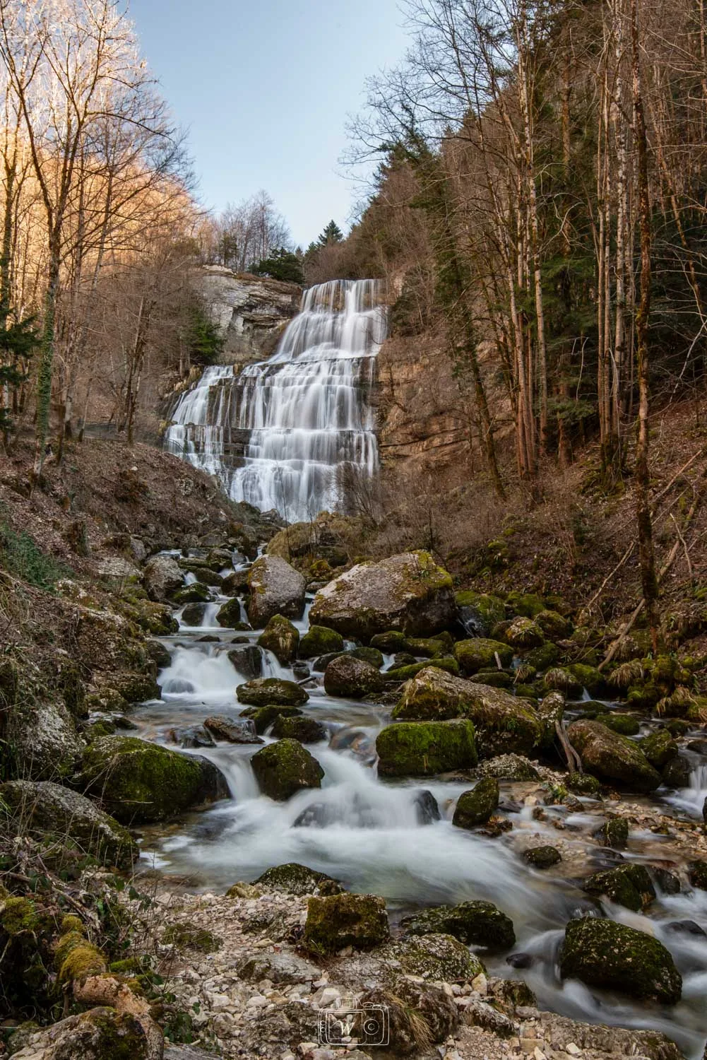 Vue verticale de la cascade de l'Éventail se déployant en escalier sur une falaise calcaire, avec un torrent rapide coulant au milieu de blocs rocheux moussus et d'une forêt jurassienne.
