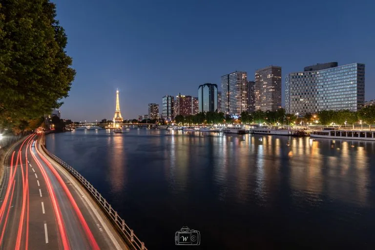 Vue nocturne de la Seine à Paris montrant les tours du quartier Beaugrenelle et la Tour Eiffel illuminée avec des traînées de phares sur la voie Georges-Pompidou.