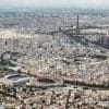 Photographie aérienne de Paris montrant le stade du Parc des Princes au premier plan et la Tour Eiffel en arrière-plan sous un ciel clair.