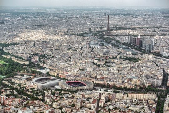Photographie aérienne de Paris montrant le stade du Parc des Princes au premier plan et la Tour Eiffel en arrière-plan sous un ciel clair.
