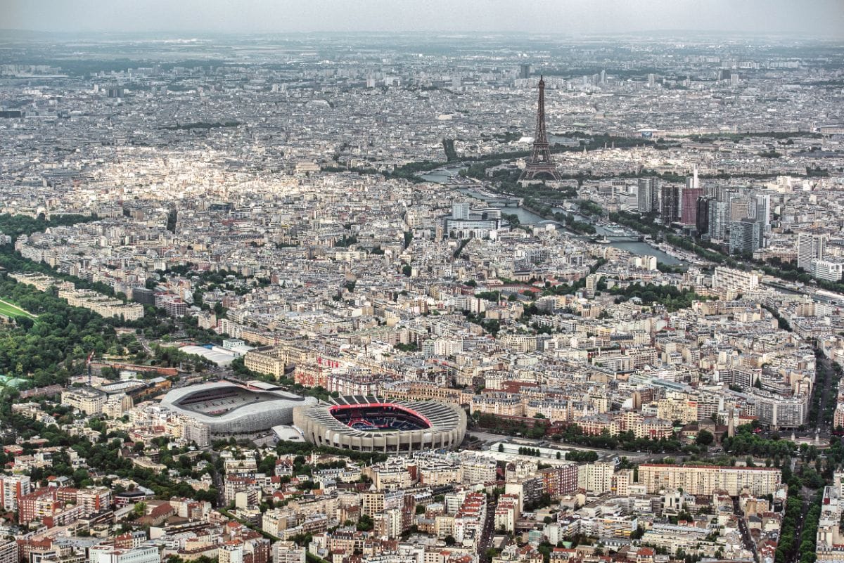 Photographie aérienne de Paris montrant le stade du Parc des Princes au premier plan et la Tour Eiffel en arrière-plan sous un ciel clair.