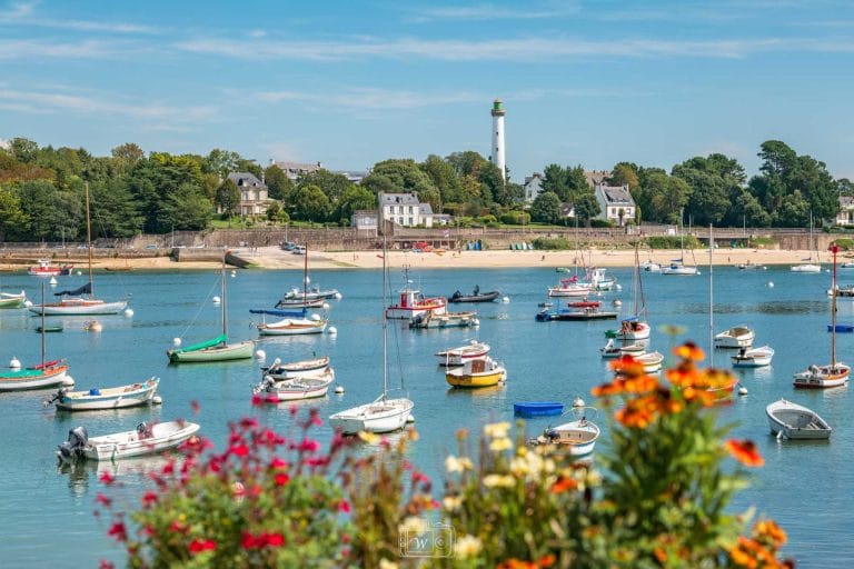 Vue sur les bateaux amarrés sur les eaux calmes de l'Odet avec le phare de la Pyramide s'élevant au-dessus des villas et de la plage de Bénodet, encadrée par des fleurs colorées au premier plan.