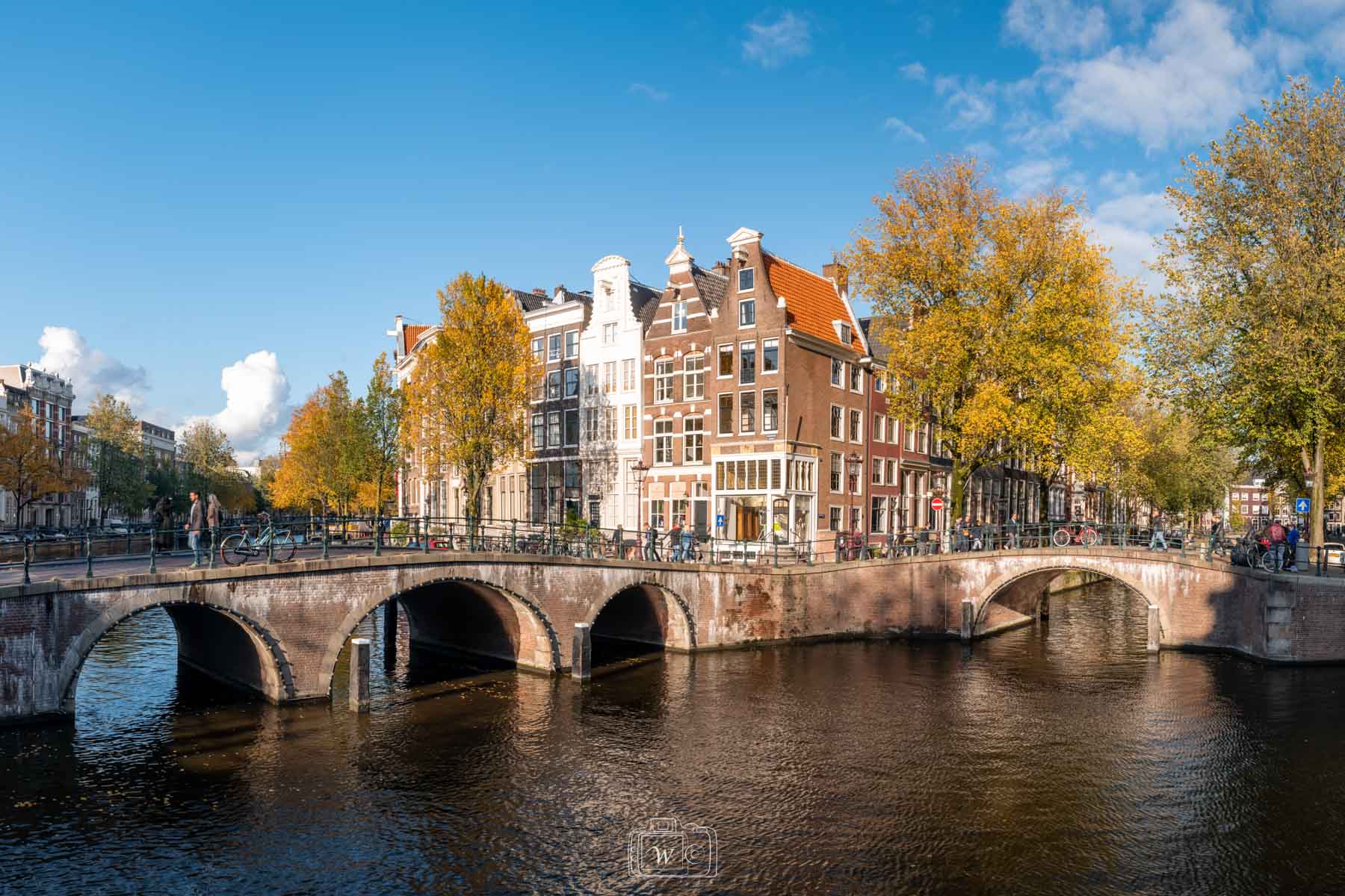 Vue d'un croisement de canaux à Amsterdam bordé de ponts voûtés en brique rouge et de maisons historiques à pignons sous les arbres dorés de l'automne.