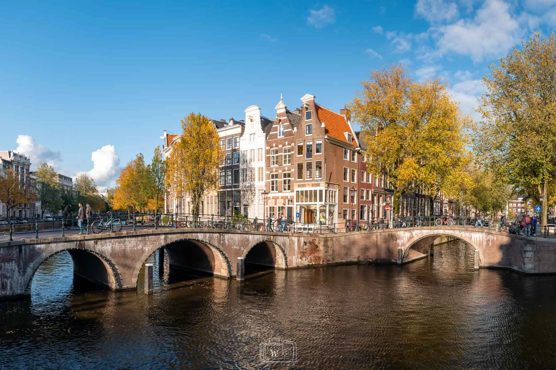 Vue d'un croisement de canaux à Amsterdam bordé de ponts voûtés en brique rouge et de maisons historiques à pignons sous les arbres dorés de l'automne.