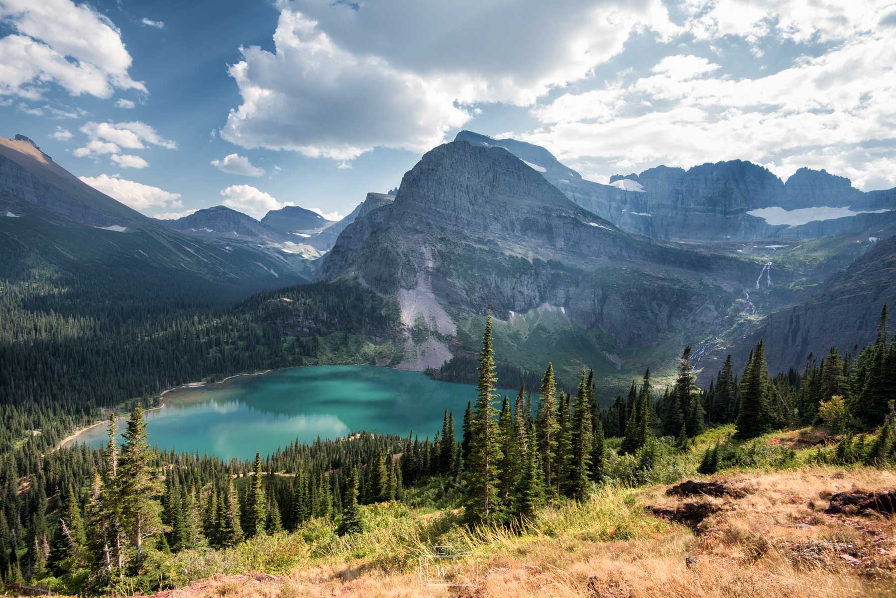 Panorama alpin sur le lac Grinnell aux eaux turquoise turquoise encaissé au milieu d'une forêt de conifères et dominé par l'imposant mont Grinnell sous un ciel nuageux dans le parc national de Glacier.